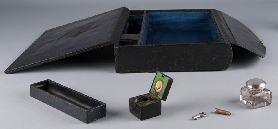 Black box with two top flaps open revealing blue interior, small black tray, two inkwells, small key, and metal ferrule on table in front of case