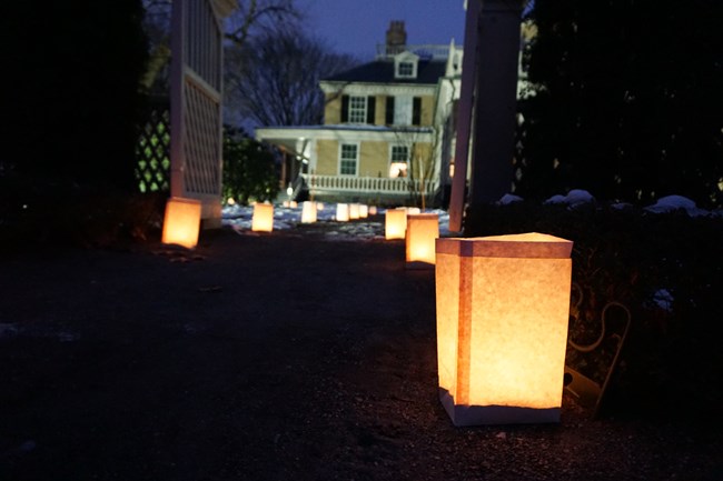 Lit paper bag luminaries at dusk with historic yellow house in background