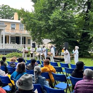 Drummers and performers wearing white, in front of a seated crowd outdoors.