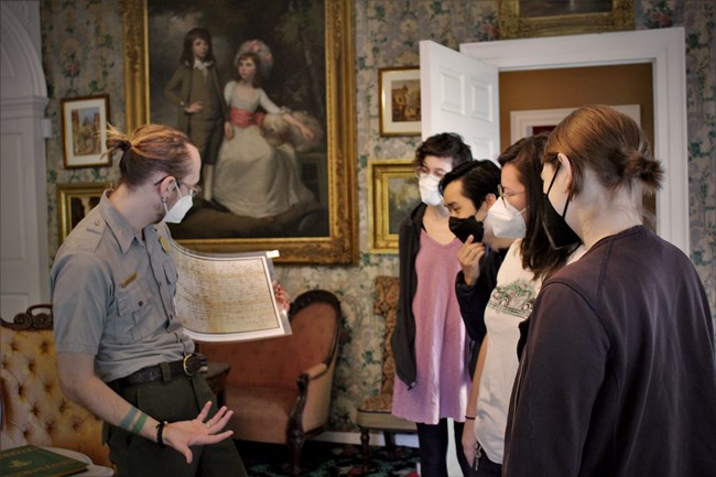 A ranger shows a handwritten historic document to four visitors in the Longfellow House parlor