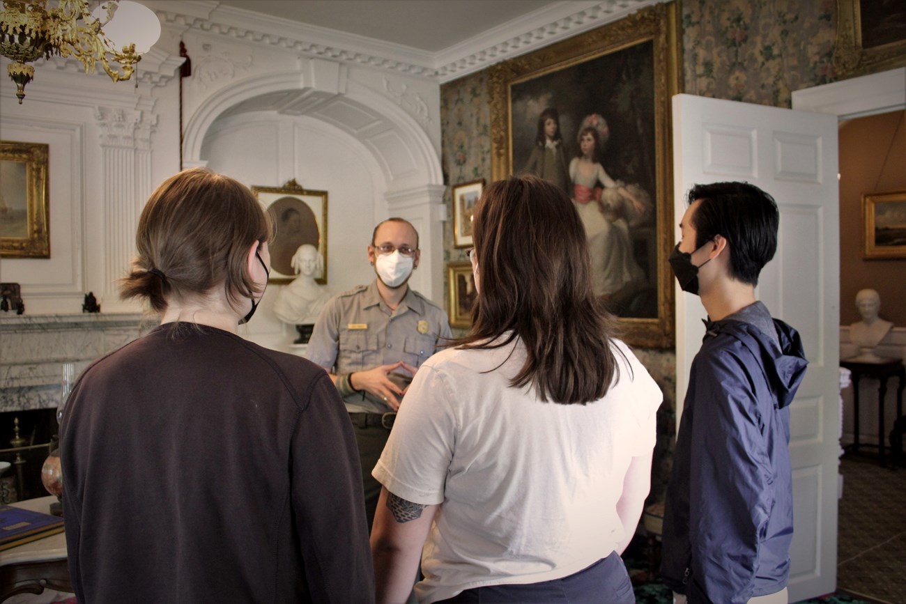 Three visitors looking away from the camera face a ranger in the Longfellow House parlor