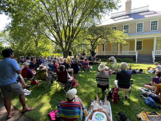 Crowd on a shaded lawn listens to and applauds musicians