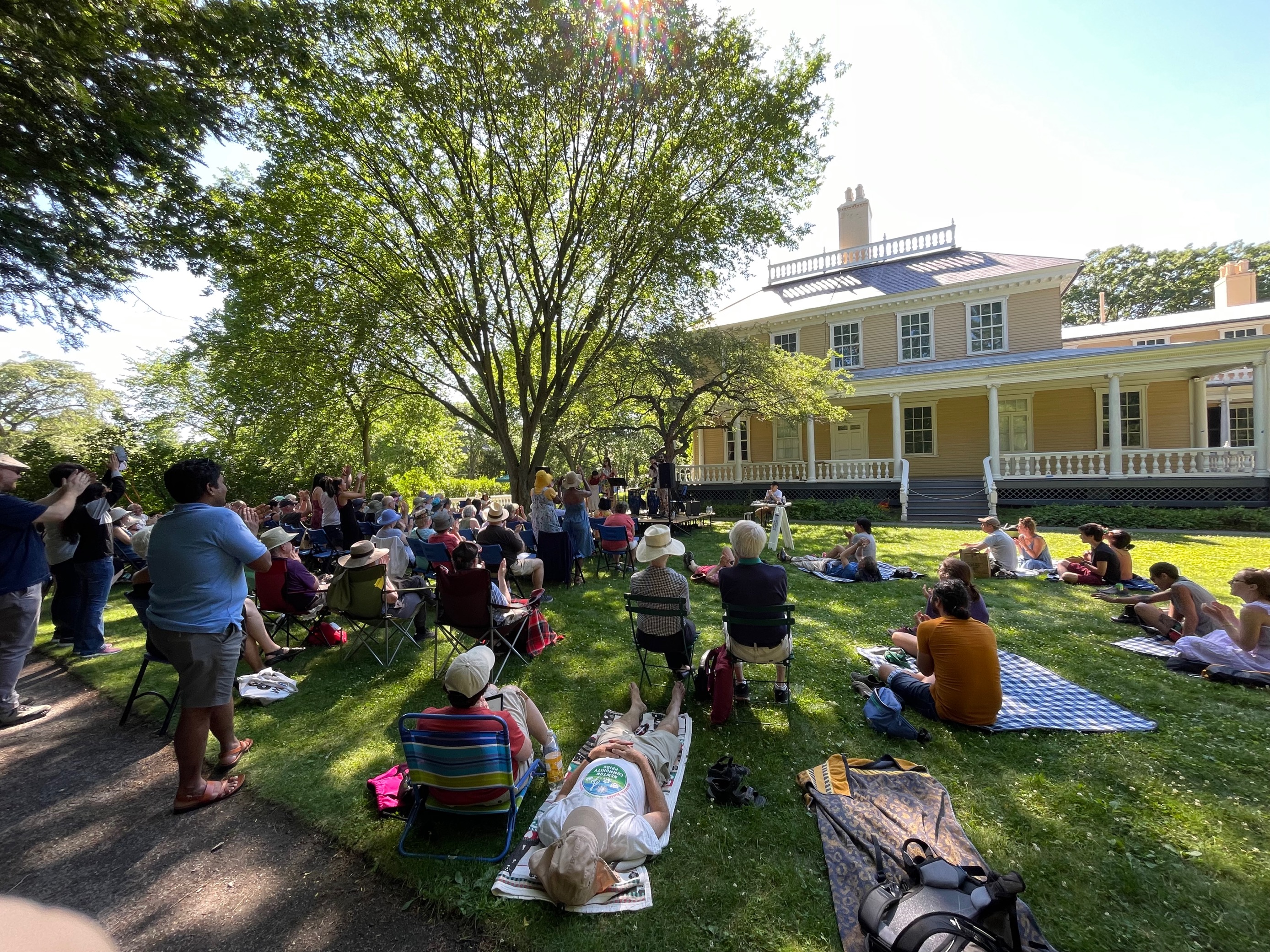 Crowd on a shaded lawn listens to and applauds musicians