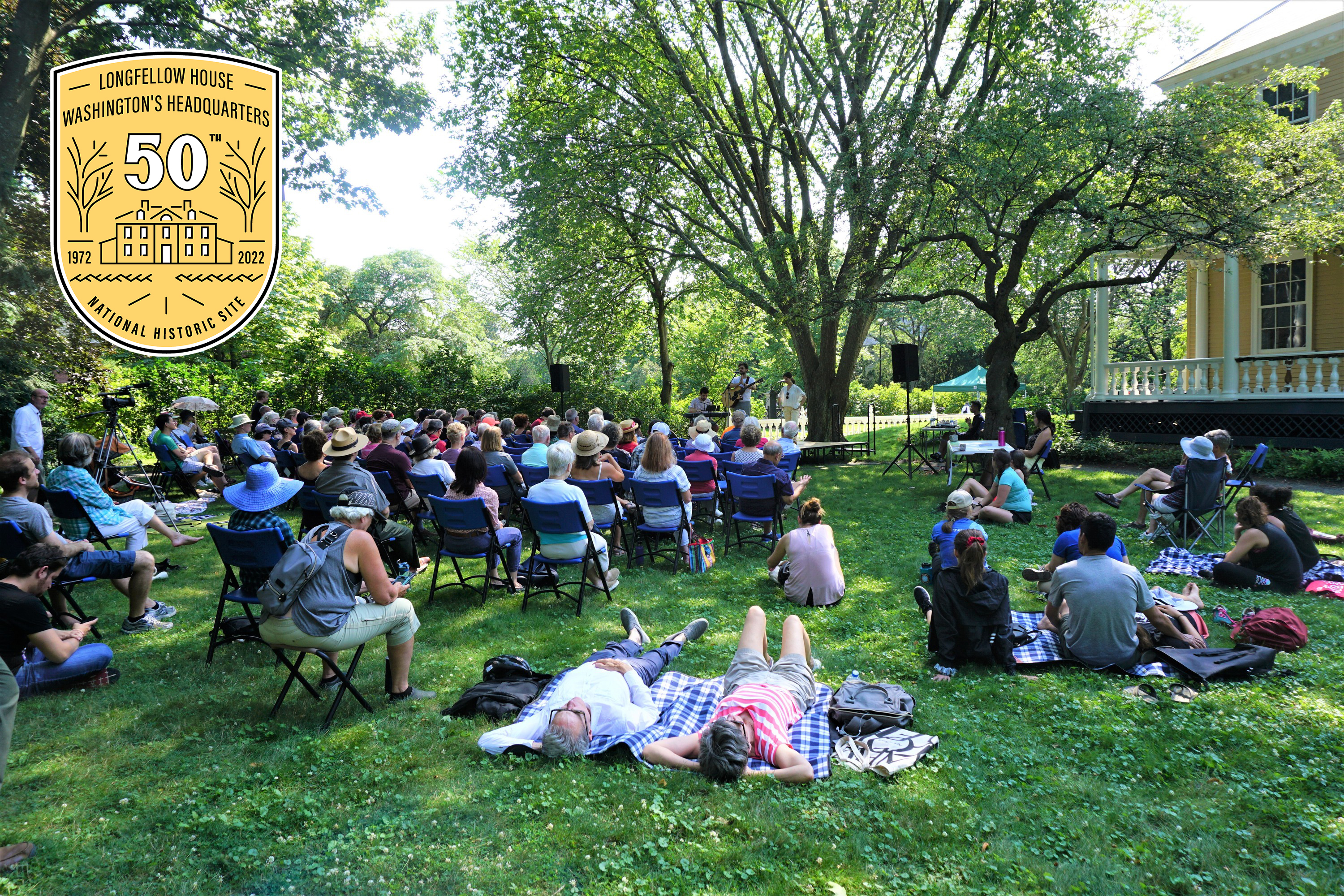 Summer Festival 2022 Crowd seated on chairs and blankets on lawn to watch concert, with 50th anniversary logo in top left corner