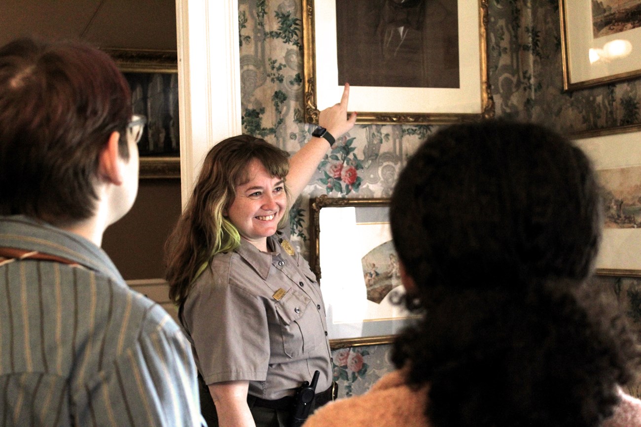 Smiling ranger points to artwork on the wall, while two visitors facing her look on