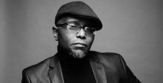 Black and white portrait of Tyehimba Jess wearing a cap, turtleneck, and blazer