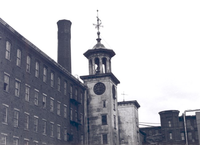 Connected to the mill building is the clock tower. The tower's exterior wall and tower is weathered. There is a bell inside the tower, with a weather vane atop. A large chimney looms behind the mill building.