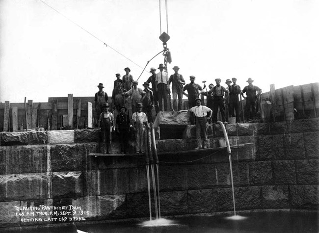 Workers setting the last cap stone for Pawtucket Dam. They pose as water trickles down the granite structure.
