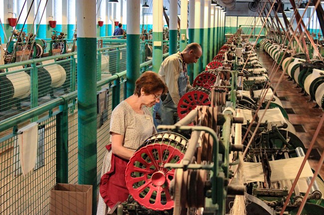 A woman and man are demonstrating weaving in a room full of power looms.