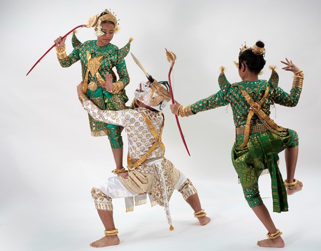 A dancer dressed as a white monkey dances with two archers. The monkey wields a dagger and has an archer poised on his thigh. All the dancers' costumes are heavily embroidered and sequined with gold ornaments.