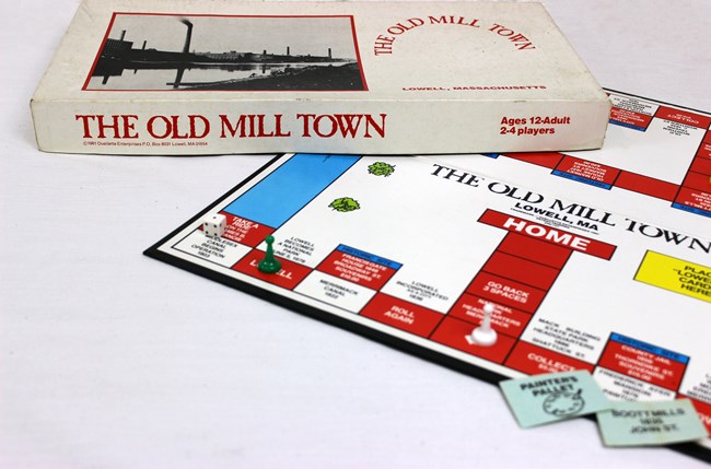 Dice, game pieces, and cards are displayed on the sites of the board game map, which comprises of square landing spaces with the names of historic sites on them. Atop the game is the box which has a photo of smokestacks and a mill overseeing the river.