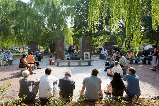 A crowd gathers around a speaker standing amidst large stone pillars in a plaza. Photos of Jack Kerouac are on the bench in front of him.