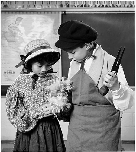 Two children, a boy and girl, are dressed as mill workers examining a wad of cotton.
