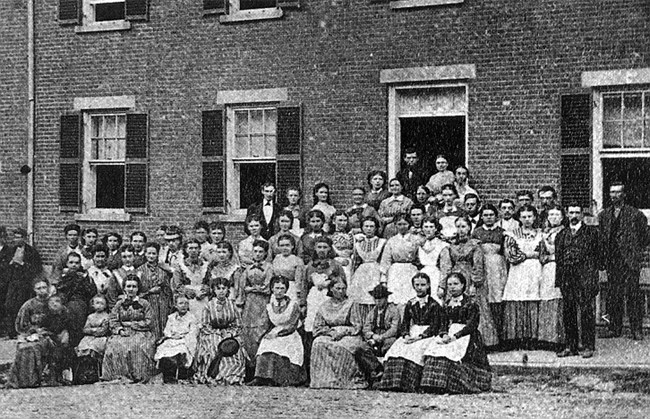 A black and white photograph of about fifty people sitting and standing in front of a two story or higher brick building. Almost all of the people in the image are women wearing dresses, and at least half are also wearing aprons.