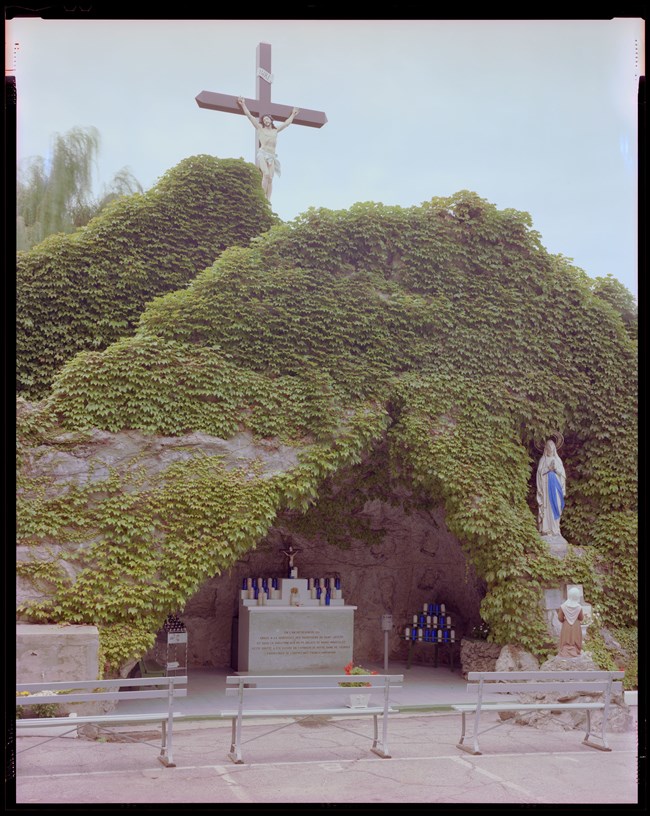 A shrine with benches station in front. The shrine is inside of a grotto with a crucifix atop the stone