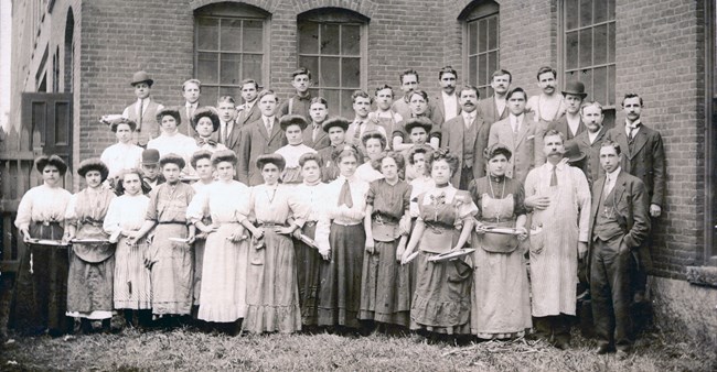 A group of workers outside Boott Mill, both men and women. Some holding loom shuttles and bobbins.