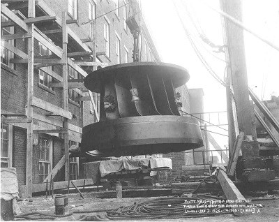A large metal water turbine is being installed in a factory in 1905