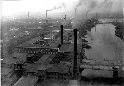 A series of smokestacks from brick mill buildings release smoke into the air