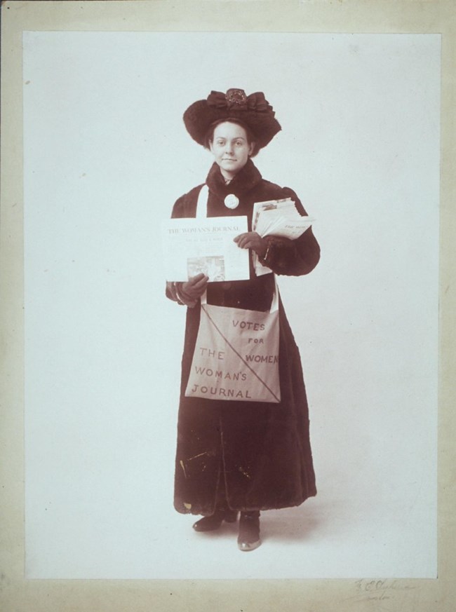 Florence Luscomb handing out copies of The Women's Journal from a bag labelled Votes for Women
