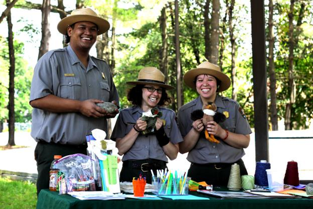 Two female park rangers and one male ranger standing a table holding two puppets and rock waiting to make crafts with kids Two female park rangers and one male ranger standing a table holding two puppets and rock waiting to make crafts with kids