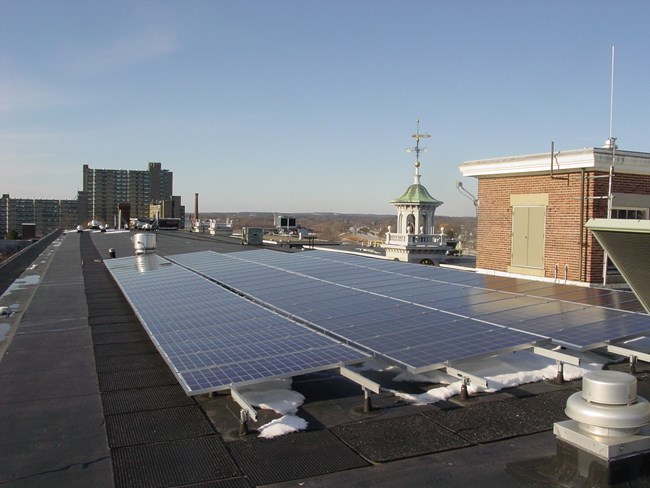 Along the roof of the Boott Museum, there are rows of solar panels lined up with the iconic Boott clocktower in the background