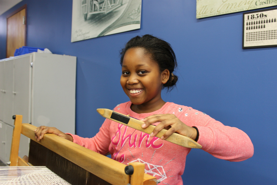 Weaving workshop. Young girl in pink sweater sits at a loom, holding a shuttle in her left hand. She has a big smile young girl working with loom, holding shuttle in left hand with smile