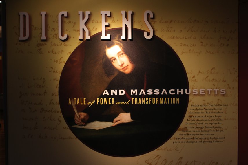 an image of Charles Dickens in the center. the word "Dickens" appears in the upper left hand corner. The image is of a painting of Dickens in his early 20's. He has black hair and holds a pen in his right hand as he looks up from a document he is writing.