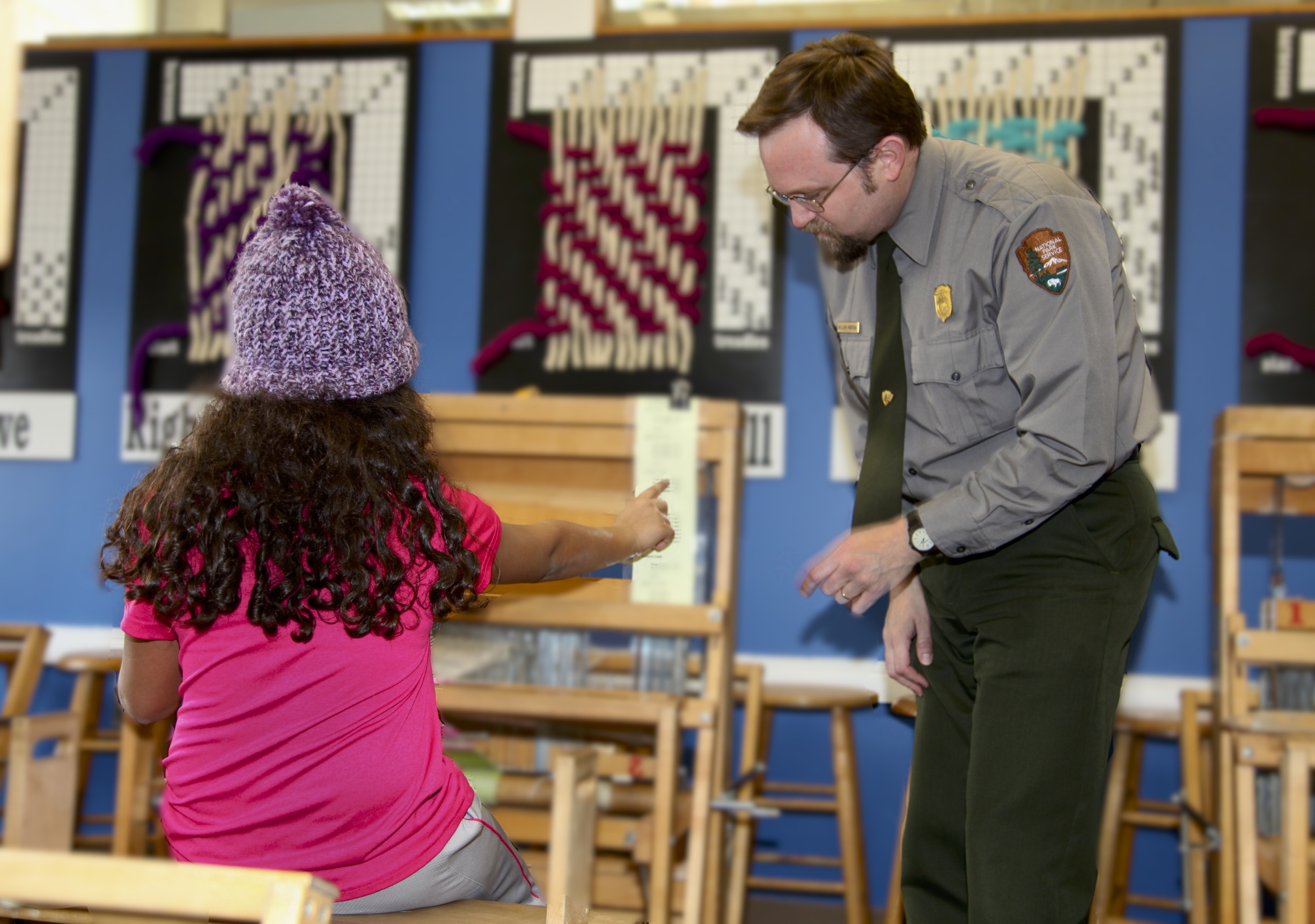 Young girl in pink shirt with a purple knit hat sits with her back to us at a small hand operated loom. To her right is a male park ranger, looking at where she is pointing.