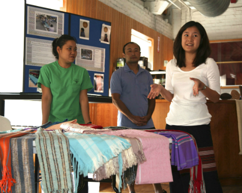Young woman in a white shirt stands to the right of a table of cloth and textiles while an older man and woman look on.