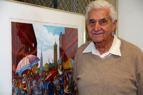 A gentleman with white hair a beige sweater and a cream color shirt look back at us with a smile on his face. To his right, an original painting of his of the first Lowell Folk Festival in 1987.