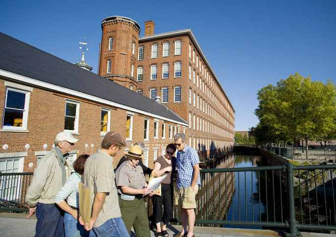 People stand around a female park ranger and behind them is a view of the Boott Cotton Mills Museum and a canal full of water