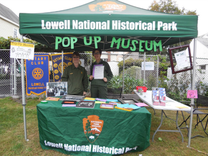 Two park rangers staff a green tent. Lowell National Historical Park appear on the top of the canopy and the words "Pop-up Museum" appear below. A table has the National Park Service arrowhead logo on it with brochures on the table.