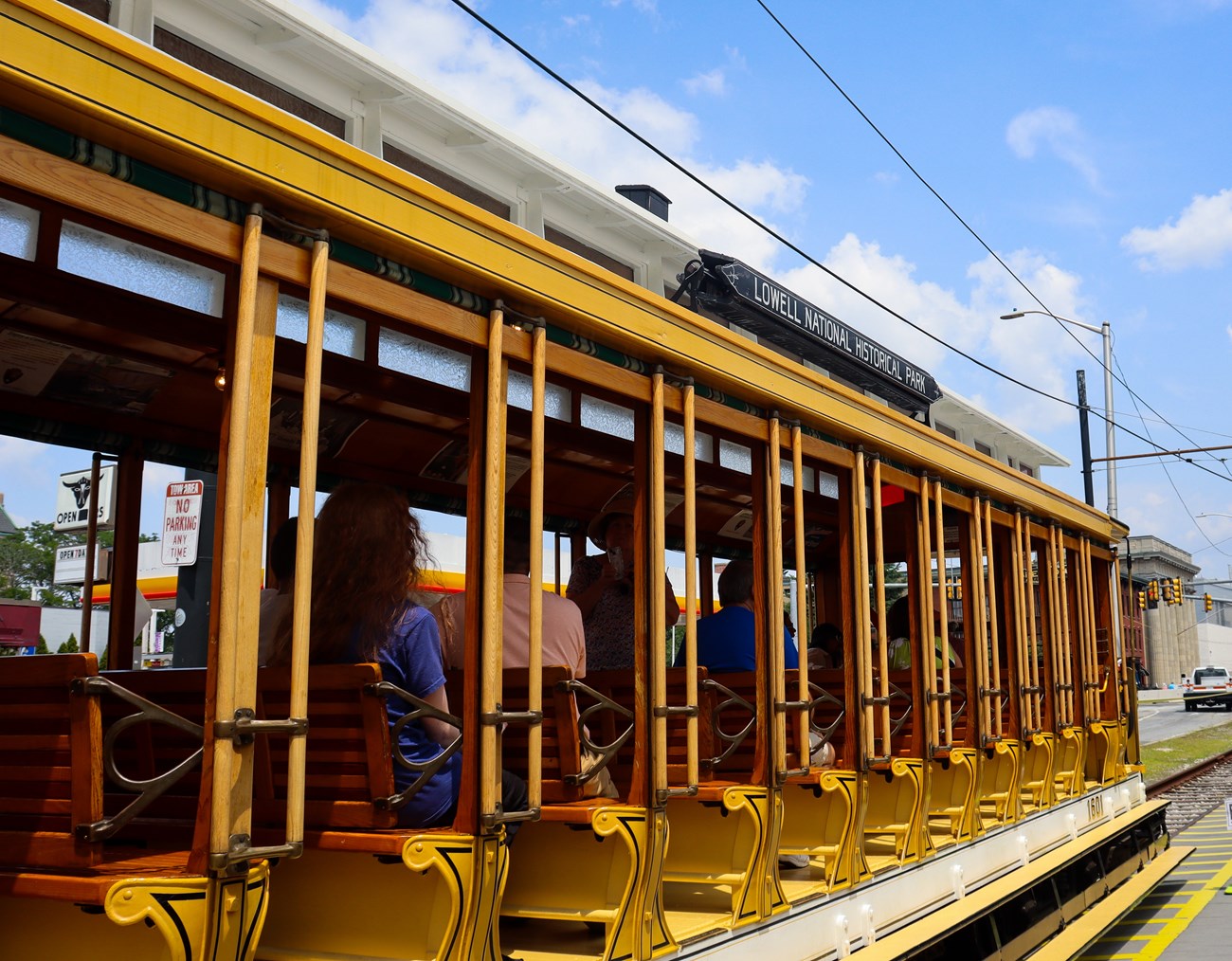 A yellow, open air trolley sits on the tracks as people sit, waiting to move. A sign on top of the trolley says "Lowell National Historical Park."