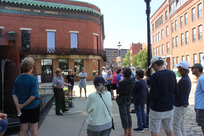 A group of visitors group around a ranger who is speaking surrounding by tall brick buildings along a street.