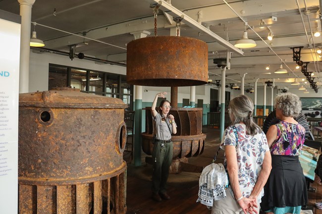 Two visitors stand listened as a ranger speaks, gesturing to large metal machinery pieces surrounding them.