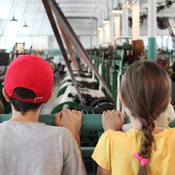 A boy and a girl with their backs to us face look into a room with machines making fabric