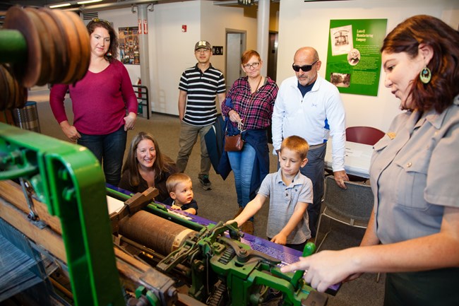 A park ranger gives a talk at a historic machine to a group of visitors of all ages