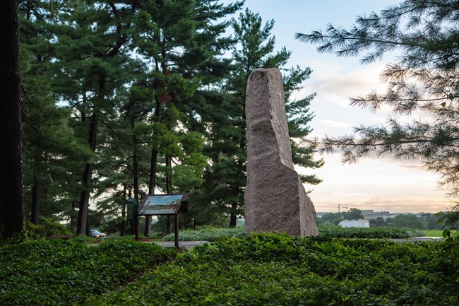 A large uncut rock sculpture rises above leafy green landscaping with a informational sign on a paved area to the left of the monolith.