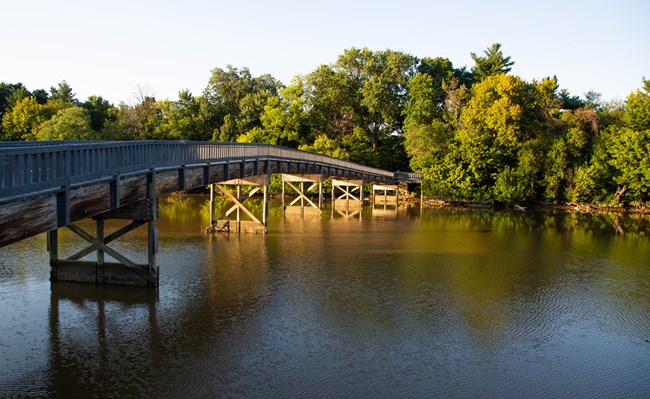 A wooden footbridge arches over a waterway with lush green trees on the opposite bank.