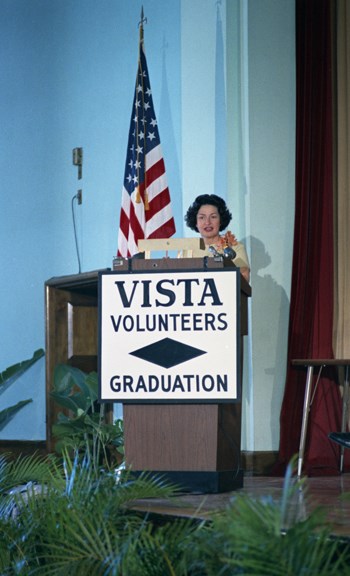 Lady Bird Johnson stands behind a lectern which displays a sign that reads "VISTA volunteers graduation."