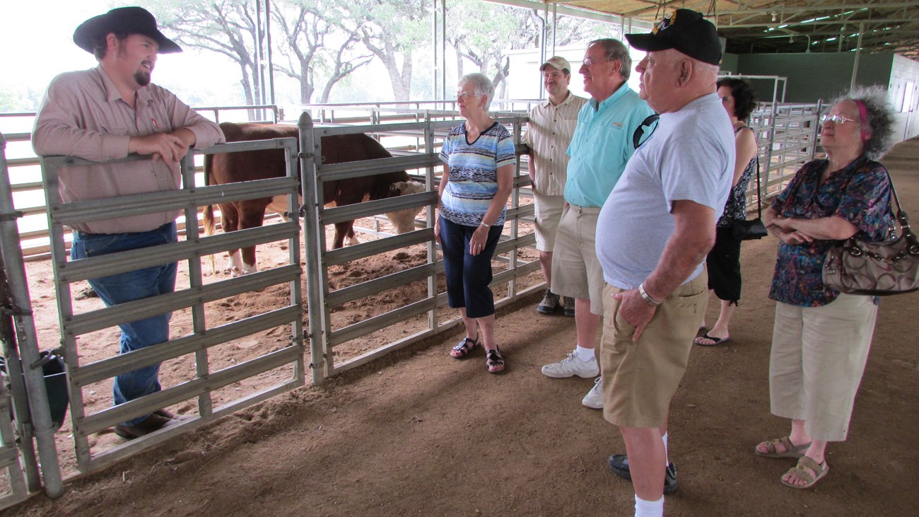 A man in cowboy hat talks to a group of people in a barn as a cow stands in the background.