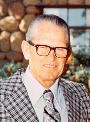 An older gentleman with thinning, gray hair wears a checkered jacket and squints behind his glasses.