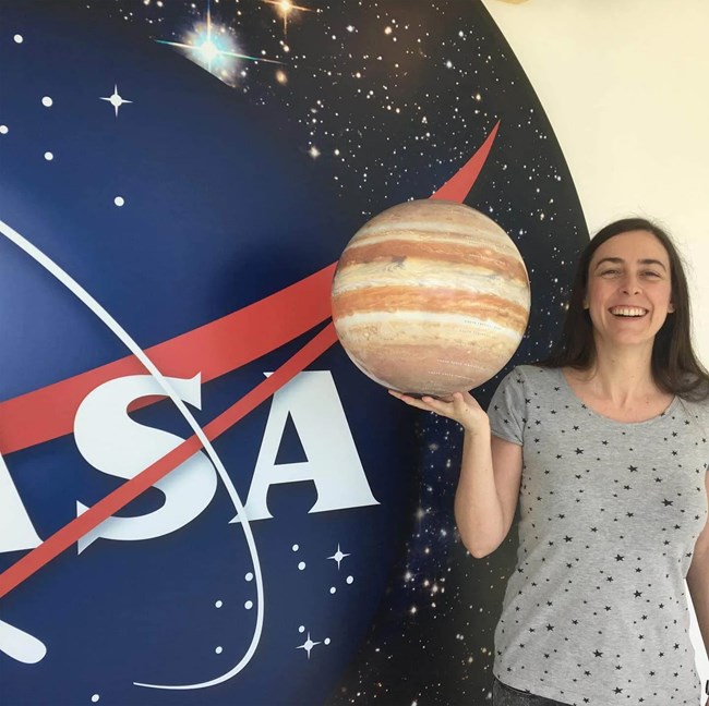 A woman holds a model of a planet while standing in front of the NASA logo.