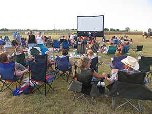 Join us for a movie under the stars! Visitors sitting in lawn chairs enjoying a movie on an outdoor screen.