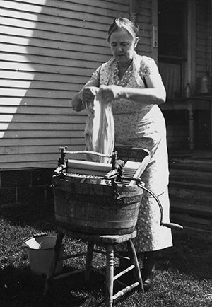 A woman stands holding a wet shirt over a tub with a washboard and hand-crank wringer. A bucket of water is on the ground beside her.