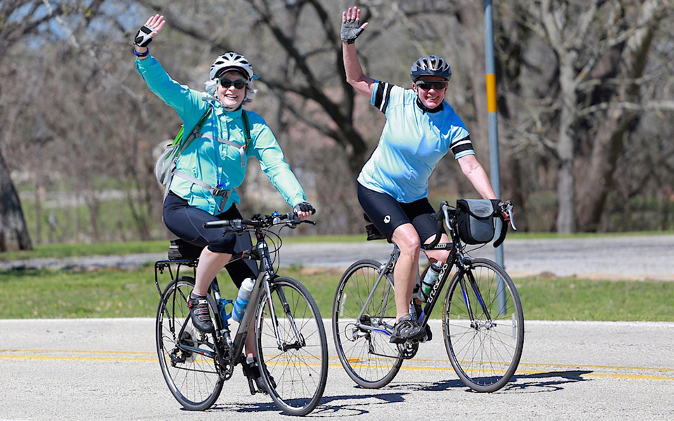 Join us for the LBJ 100 on March 30, 2019! Two bicycle riders wave as they travel a paved road in the Texas Hill Country