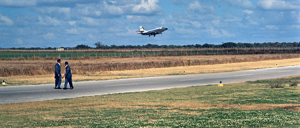 Two men in uniform stand on an asphalt strip among pastures to watch a plane in flight.