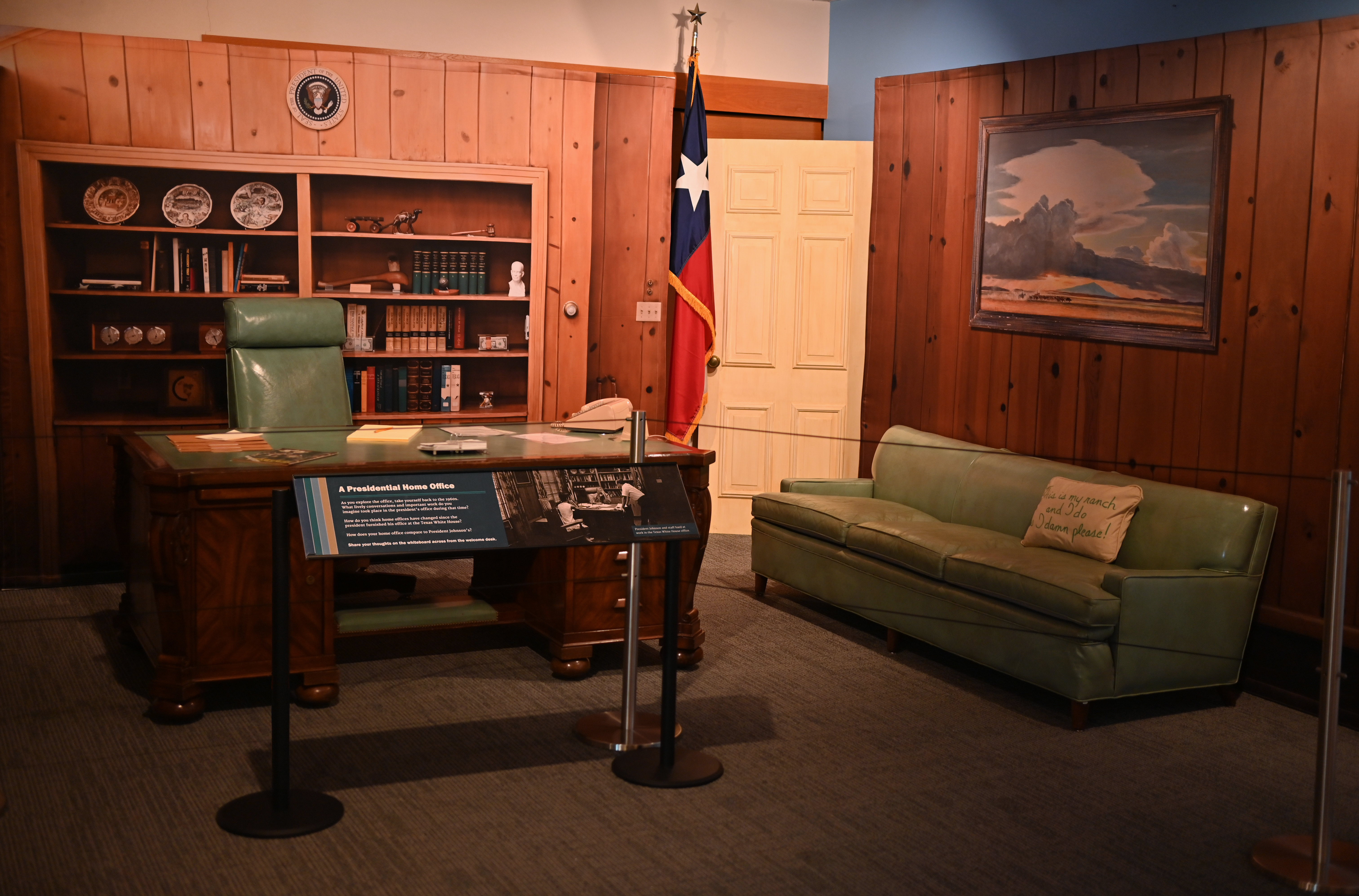 A desk and teal sofa are displayed in front of a shelf backdrop. An interpretive panel stands in front.