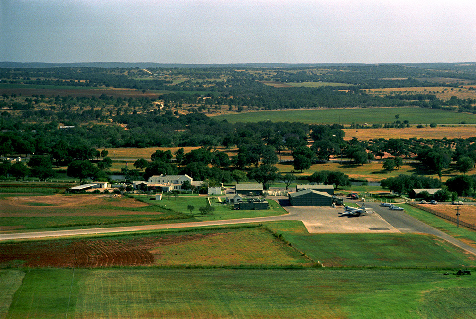 Aerial view of a large house and paved taxiway among green pastures and hills.