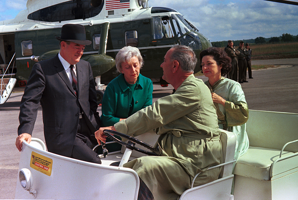President and Mrs. Johnson sit in a golf cart talking to Vice President Hubert Humphrey and his wife. A helicopter is in the background.